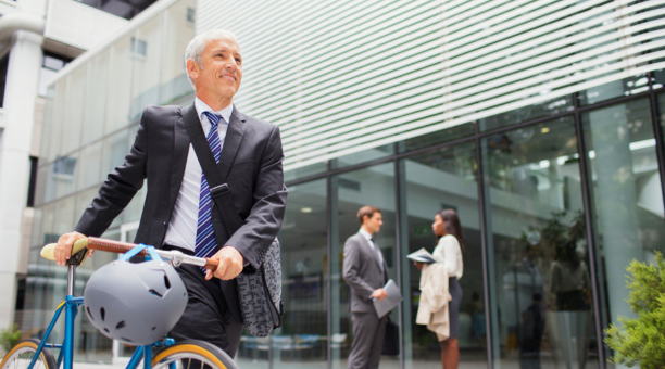 Man bikes to work with other professionals behind him in front of office building. 