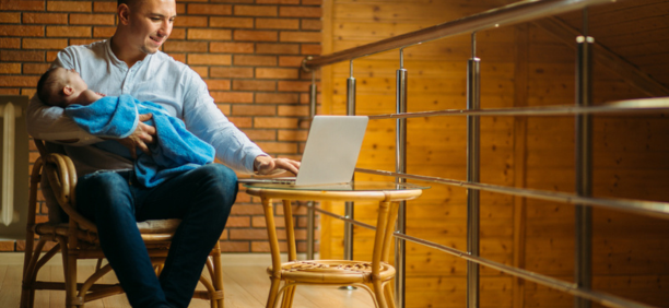 30-something man holds a sleeping baby on his lap while he works on his open laptop.