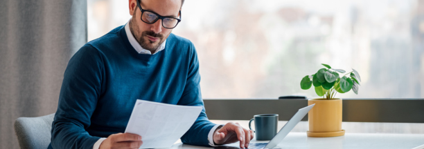 Man in blue sweater with glasses looks down at paper in his hand while sitting at a desk working. 